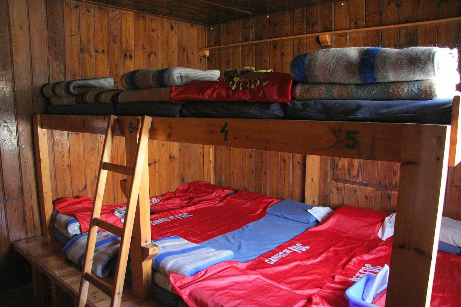 Bunk beds with blankets and red sleeping bags inside a rustic wooden refuge room at Refugi d'Estany Llong.