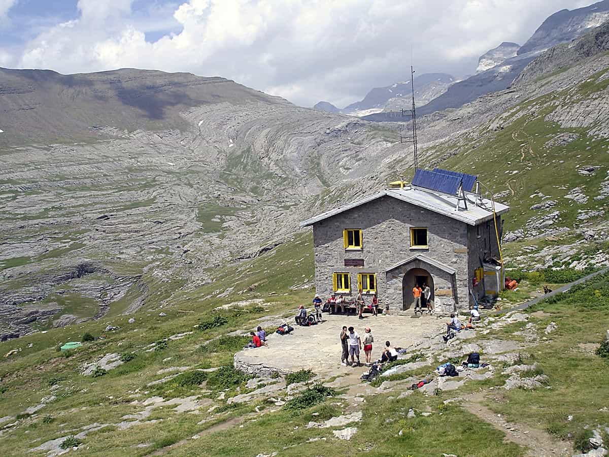 Refugio de Góriz stone mountain hut with hikers resting outside in grassy alpine terrain.