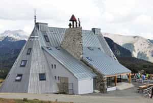 Modern mountain refuge building with metal roof and stone chimney against a backdrop of rocky peaks.