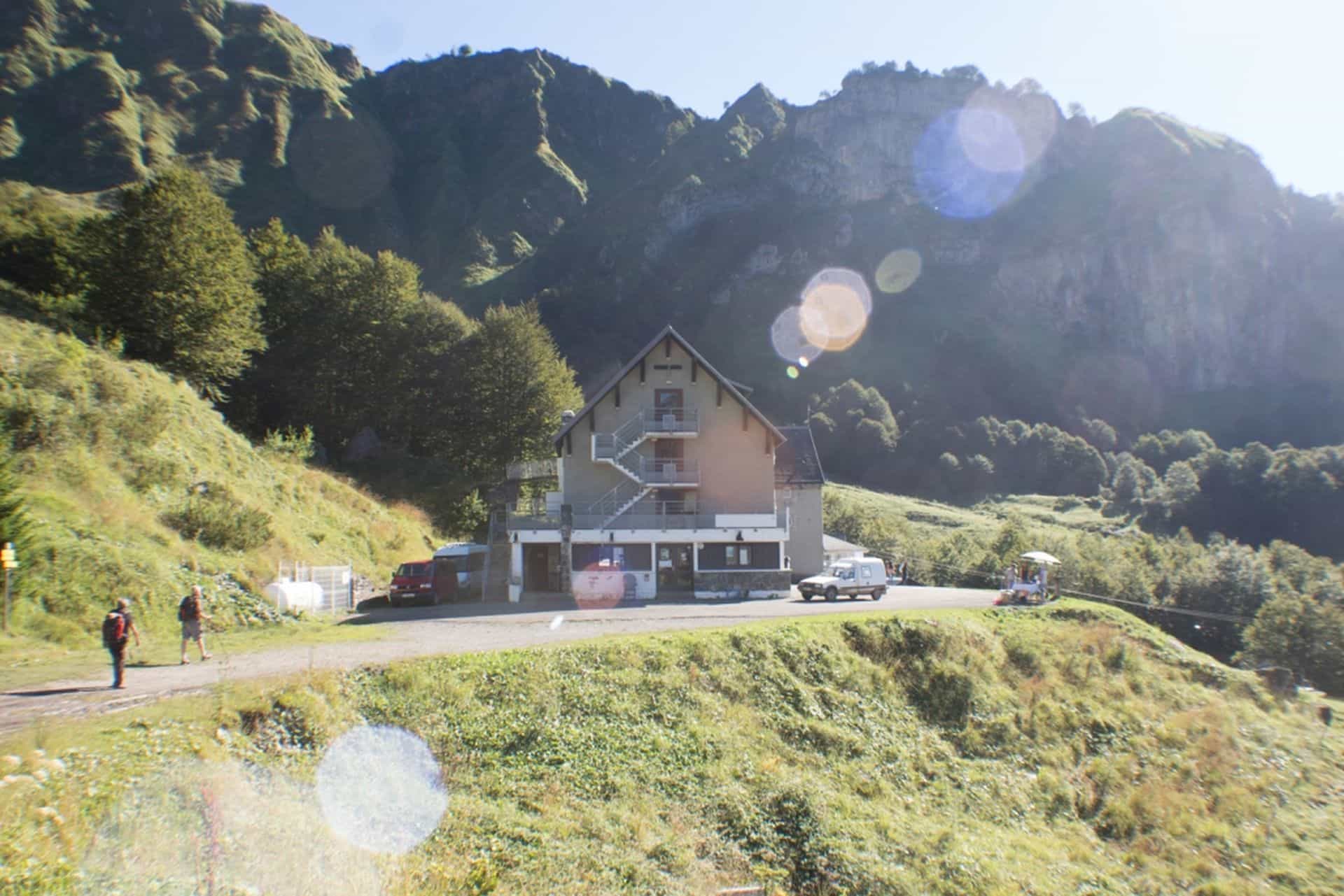 Refuge building nestled in green mountainside with hikers and parked vehicles.