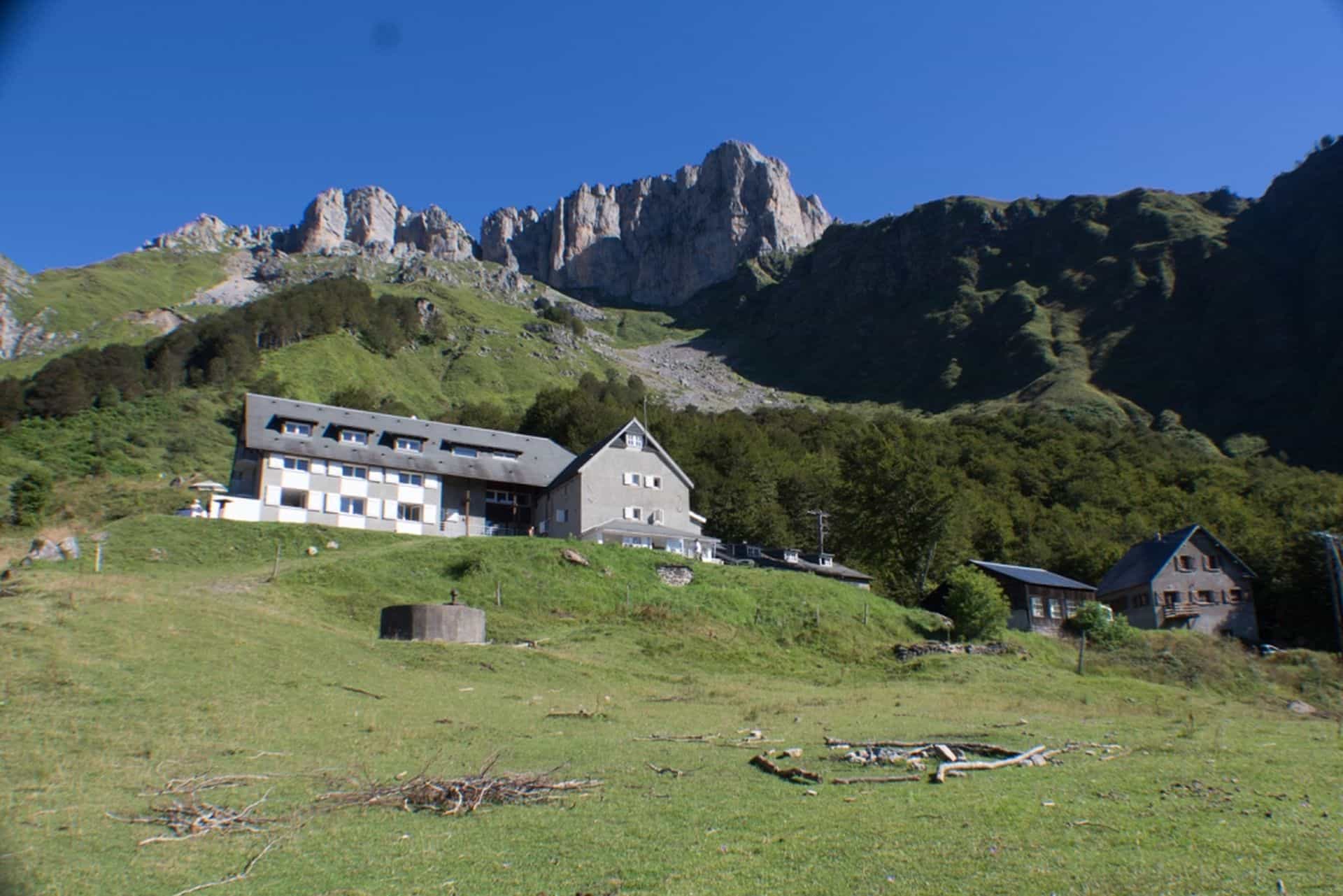 Refuge Laberouat building on grassy slope below rugged mountain peaks under clear blue sky.