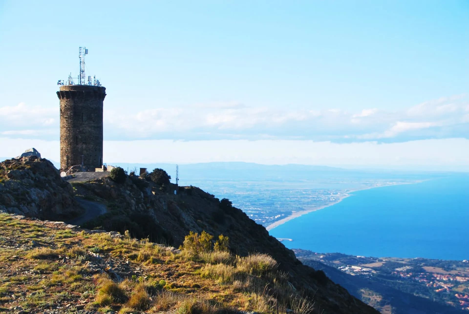 Stone tower with antennas on a grassy mountain overlooking a curved coastline and blue sea.