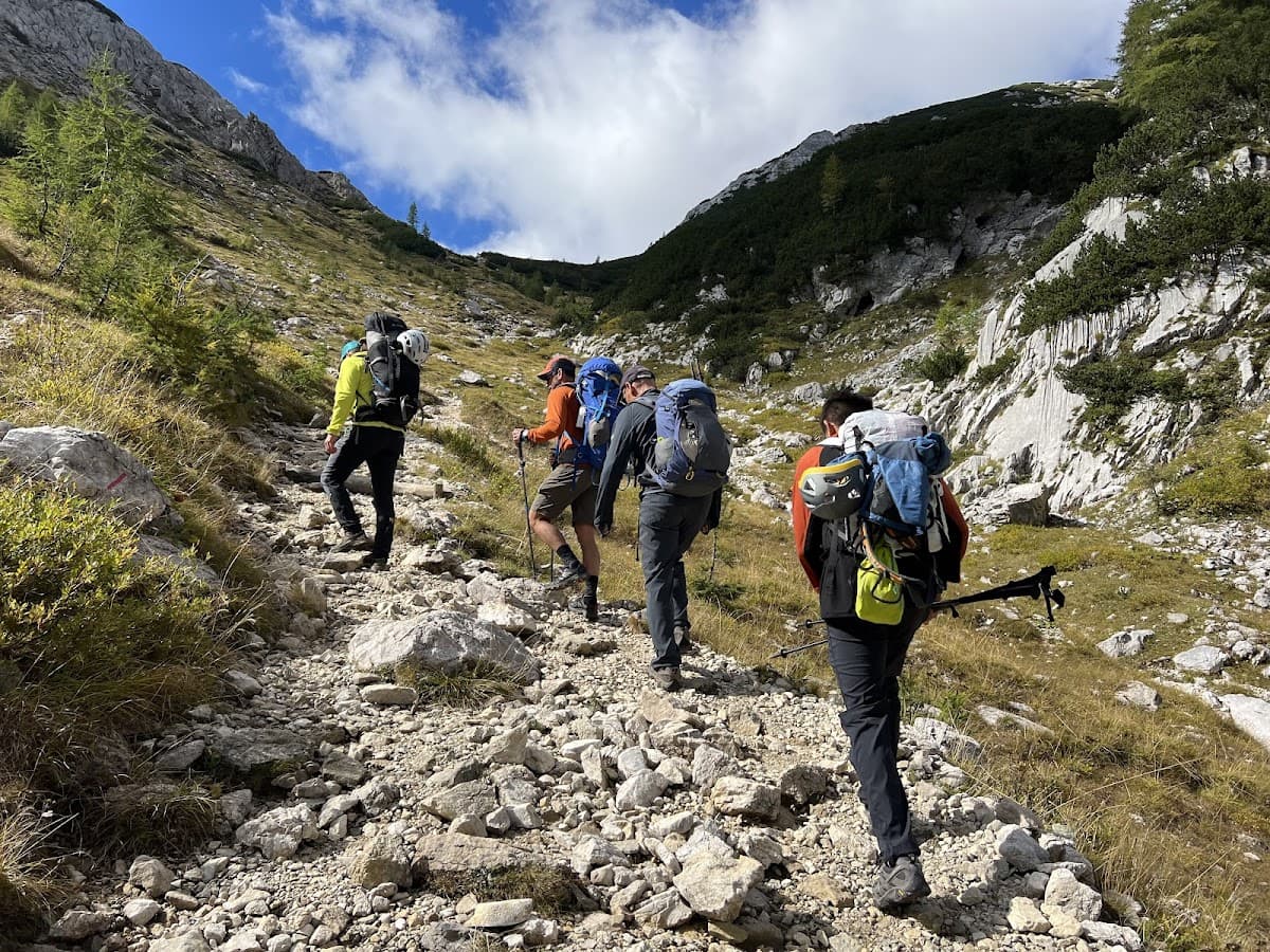 Hikers with backpacks ascend a rocky, grassy mountain trail under a partly cloudy sky.