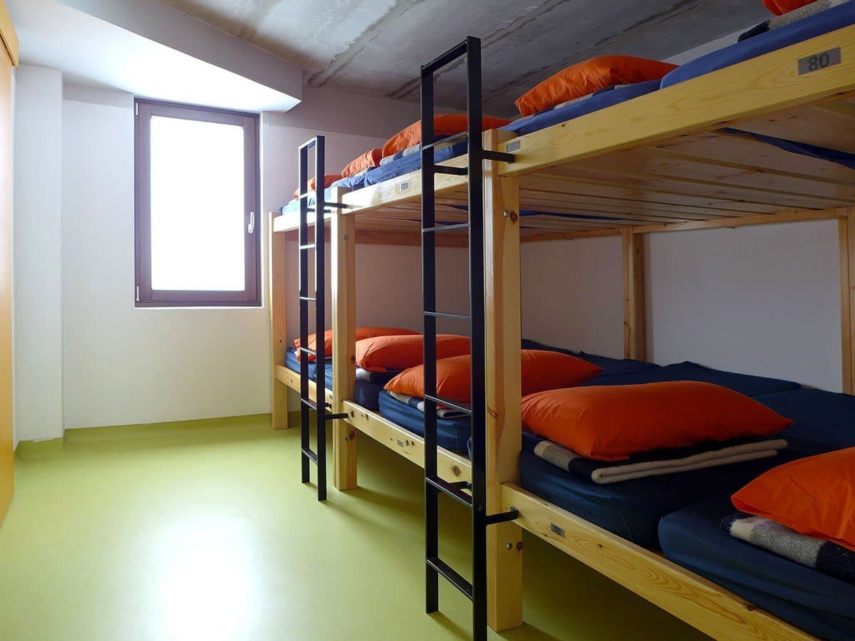 Wooden bunk beds with orange pillows in a simple dormitory room with a bright window.
