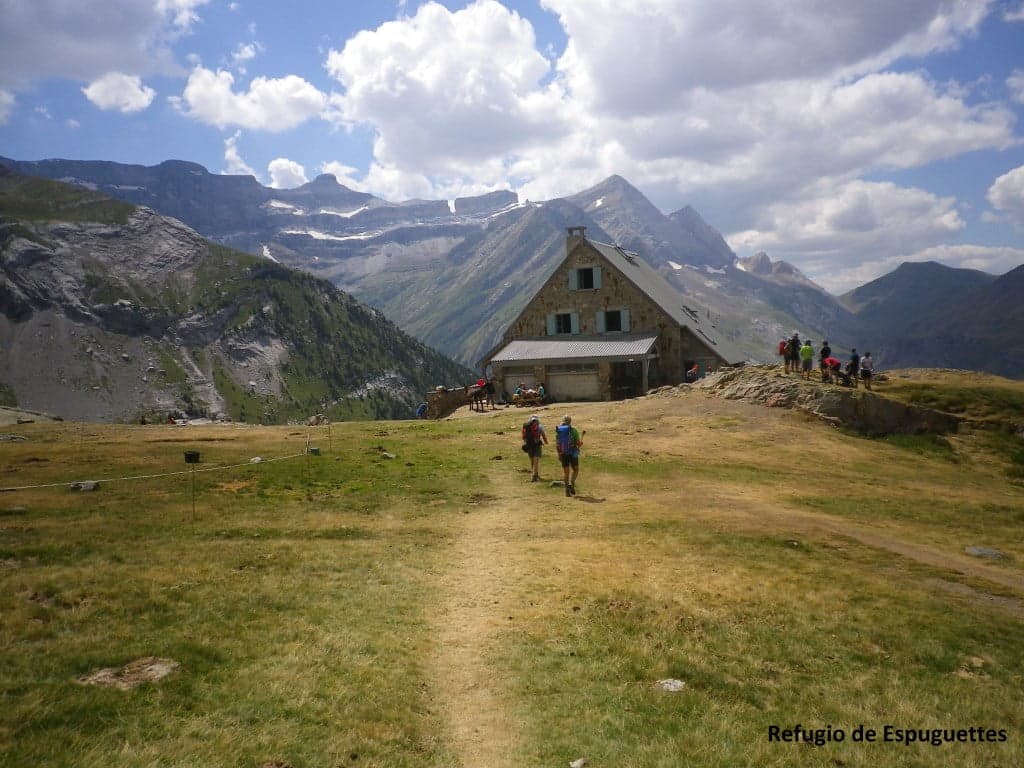 Hikers approaching Refugio de Espuguettes with snow-capped mountains under a cloudy sky.