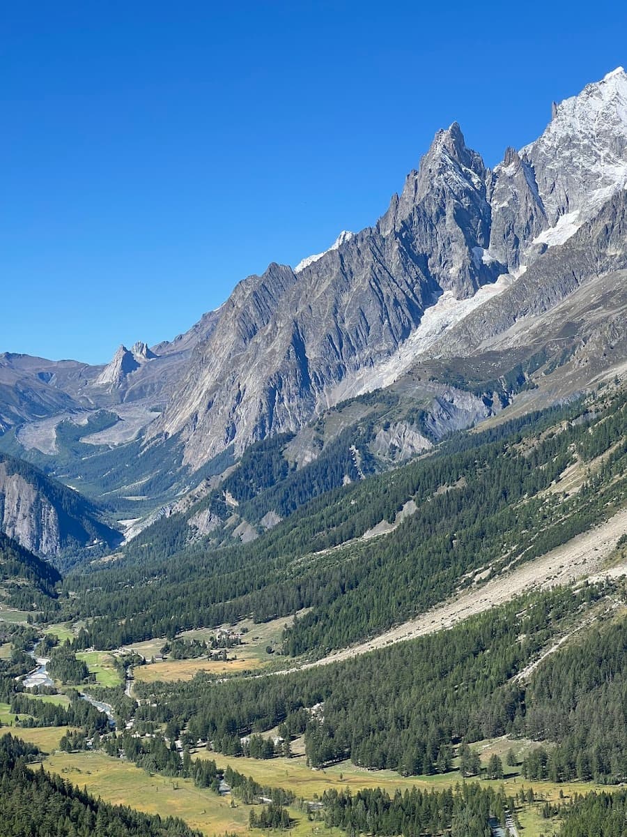 Rugged, snow-capped mountains tower over a forested valley with a river and small clearings under a clear blue sky.