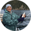 Man fishing on boat holding small fish with forested rocky coastline background