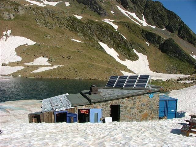 Refuge de Vénasque hut with solar panels by a lake, surrounded by snow and green mountains.