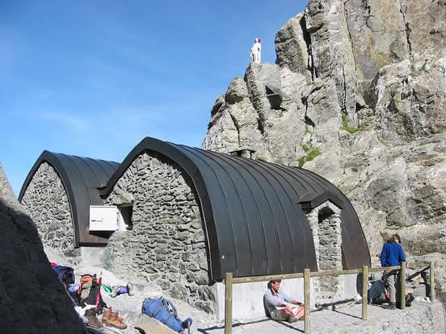 Stone mountain refuge with curved metal roof nestled against a rocky cliff face under blue sky.