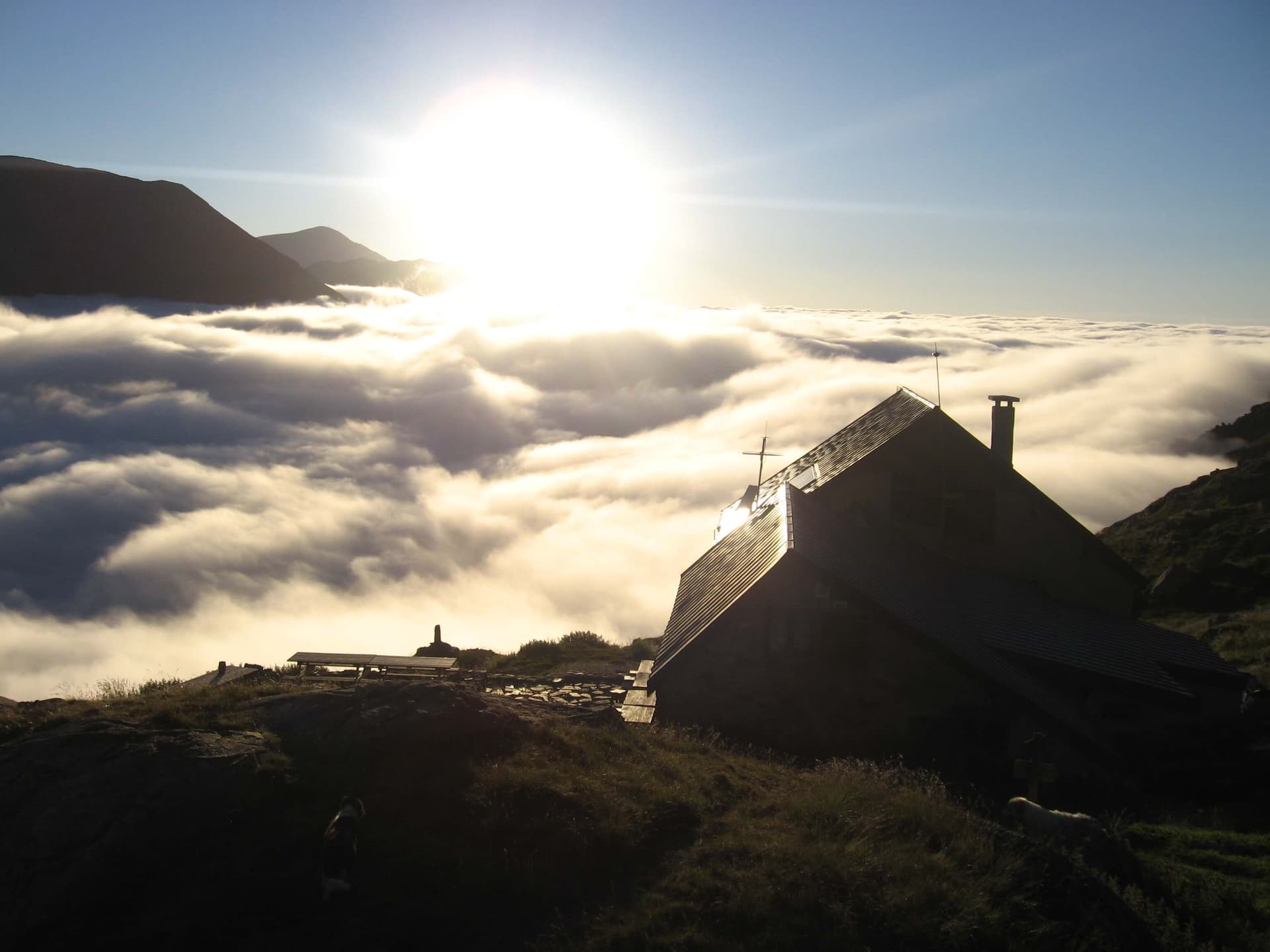 Mountain refuge above sea of clouds at sunrise with dark silhouette of building and mountains.