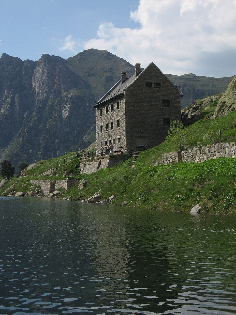 Stone mountain refuge next to dark lake with steep green slopes and rugged peaks
