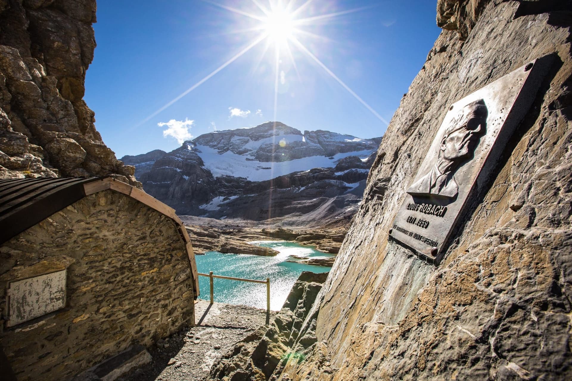 Mountain hut and Louis Robach plaque overlooking turquoise alpine lake and snow-capped peak.