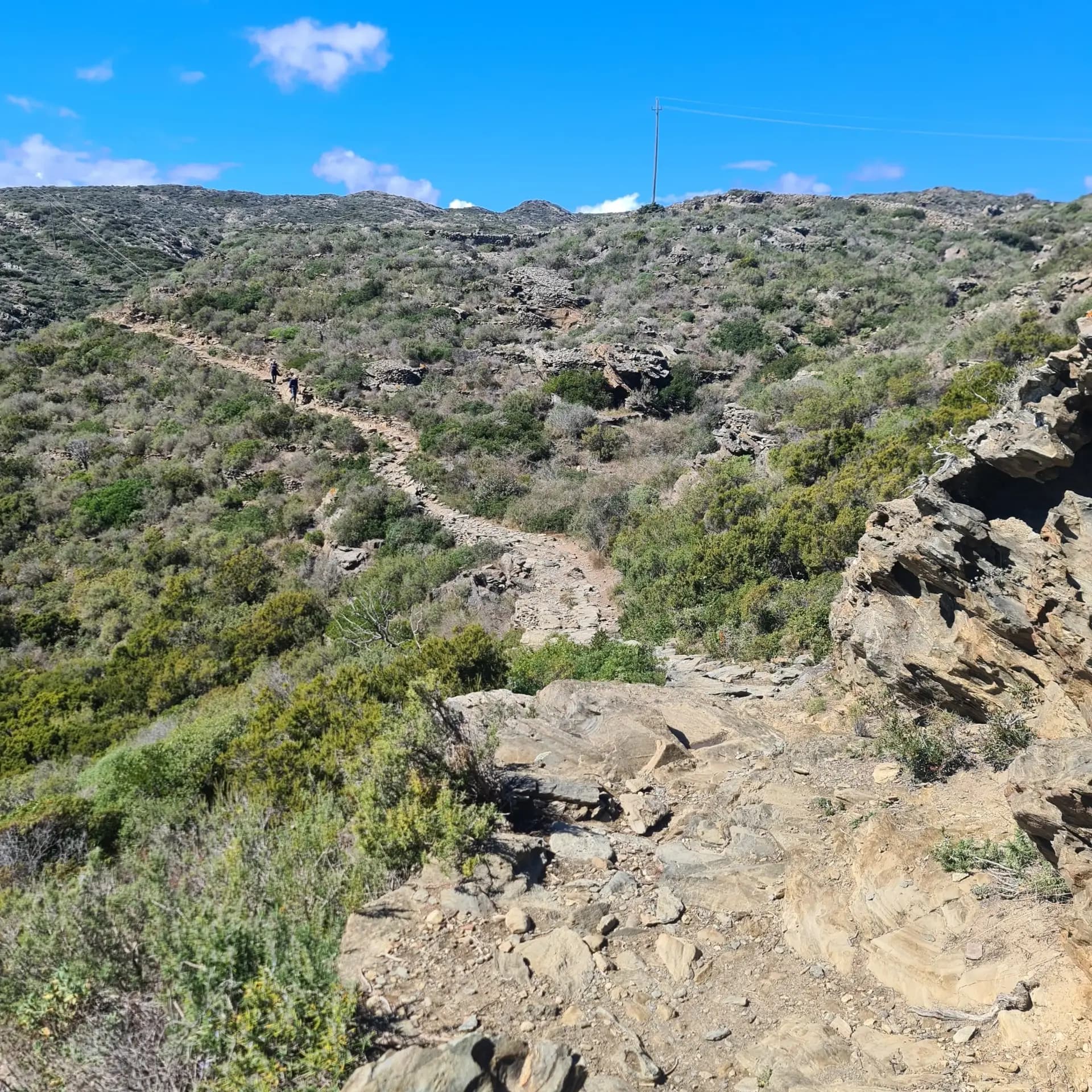 Hikers on a rocky mountain trail through a Mediterranean landscape with green shrubs under a clear blue sky.