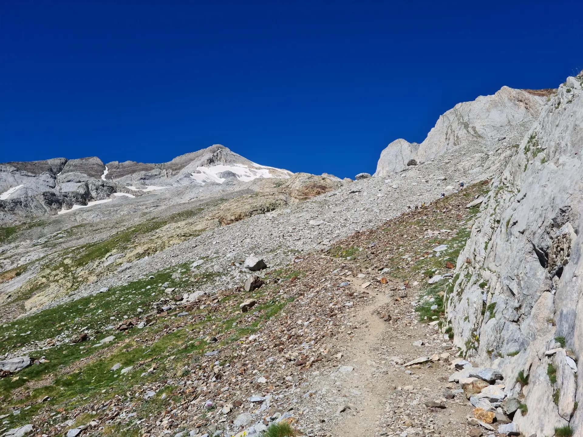 Hiking trail on a rocky mountain slope with snow-capped peaks under a clear blue sky.