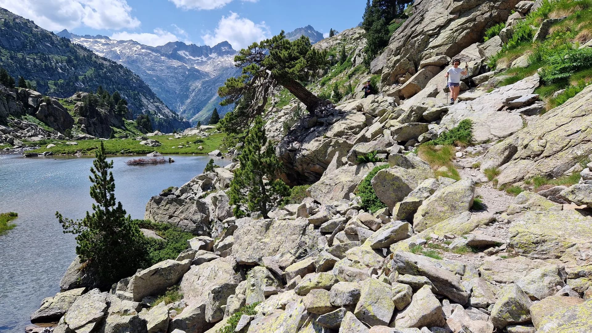 Hiker with trekking poles on a rocky trail beside a mountain lake with snow-capped peaks in the background.
