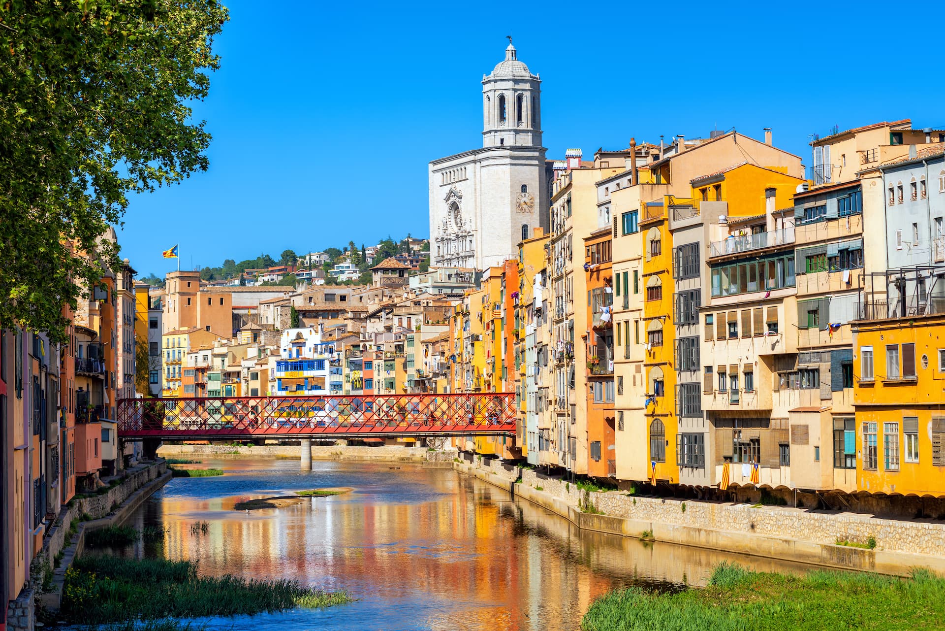Colorful buildings line a river in Girona old town with a red bridge and cathedral tower.