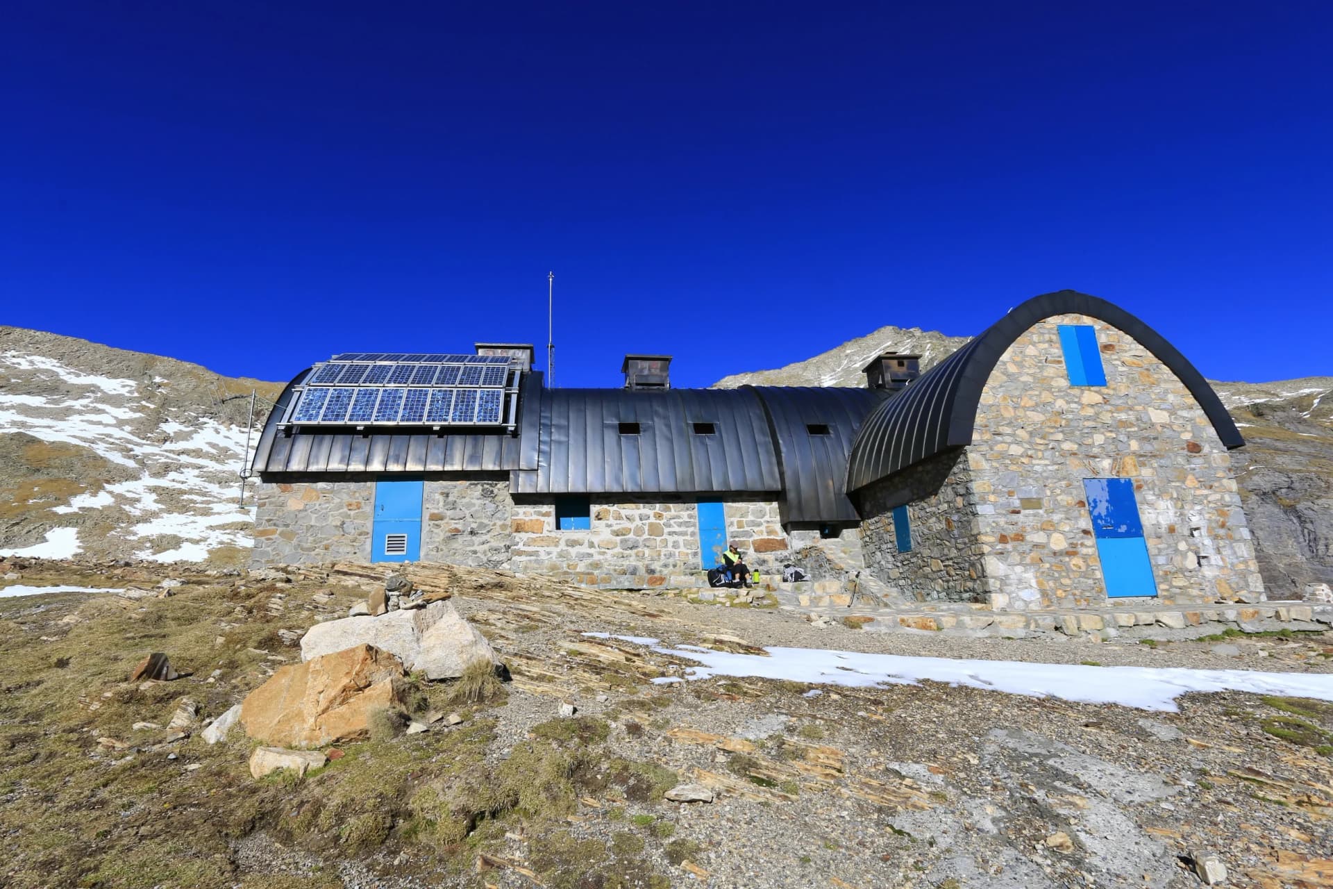 Stone refuge with solar panels and blue doors on rocky mountain slope under clear blue sky.