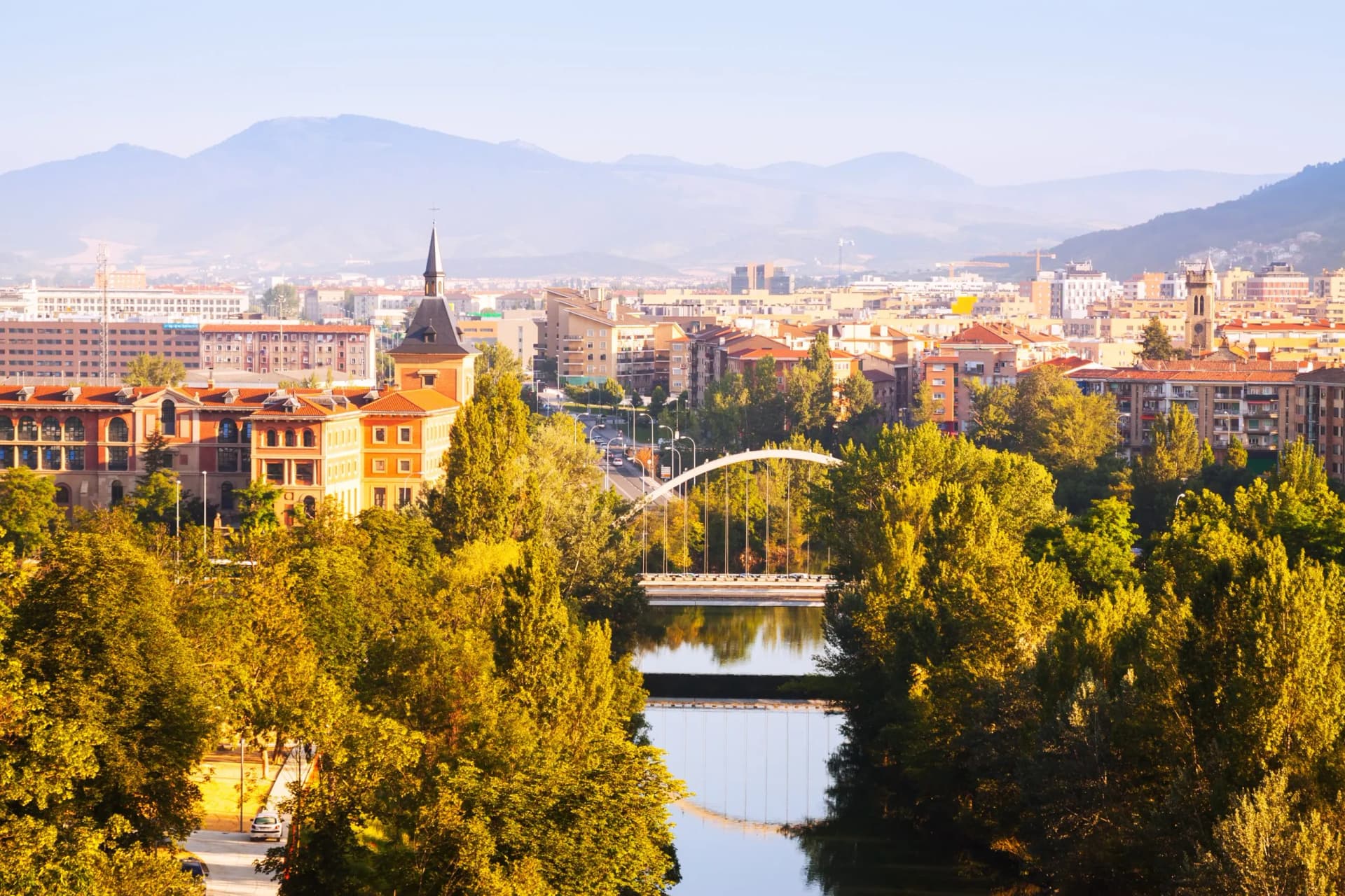 Cityscape of Pamplona with bridge over river, trees, and mountains in background.