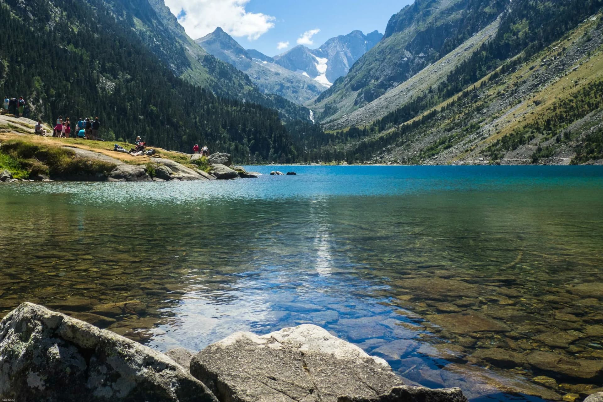 Alpine lake with clear turquoise water, rocky shore, and steep, forested mountains under a blue sky.