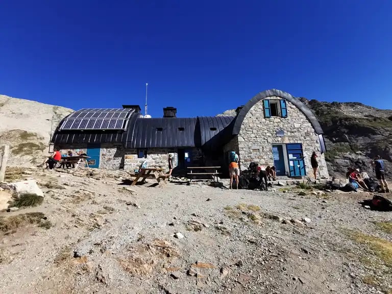 Stone mountain refuge with solar panels and hikers resting under a clear blue sky in Refuge Bayssellance.