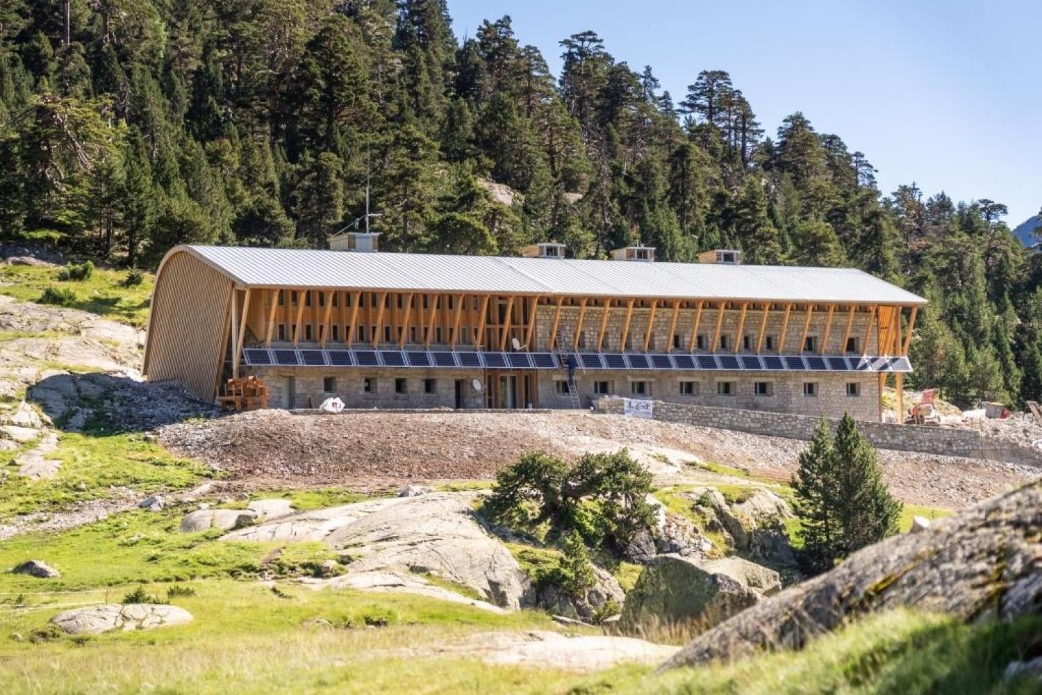 Refuge Marcadau with stone base and wood facade nestled in a sunny mountain forest setting.