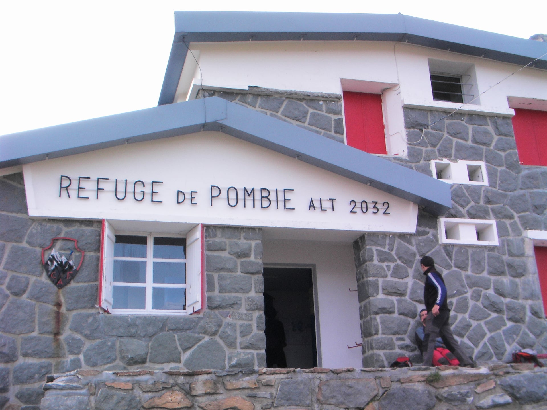 Refuge de Pombie entrance, stone building with red shutters, hikers arriving at altitude 2032.