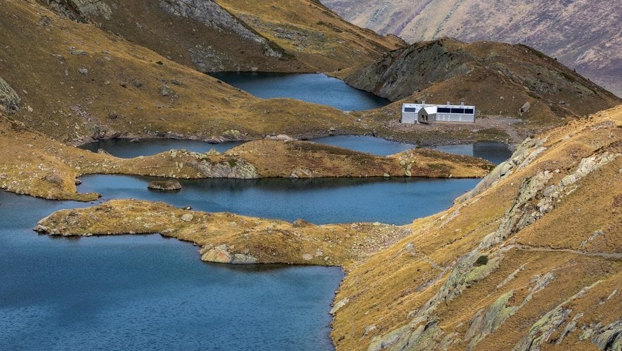 Mountain refuge building nestled between several dark blue alpine lakes surrounded by dry, grassy slopes.