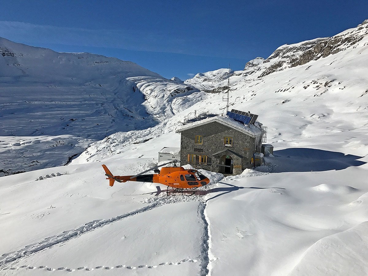 Orange helicopter landed beside stone mountain hut surrounded by deep snow under clear blue sky.