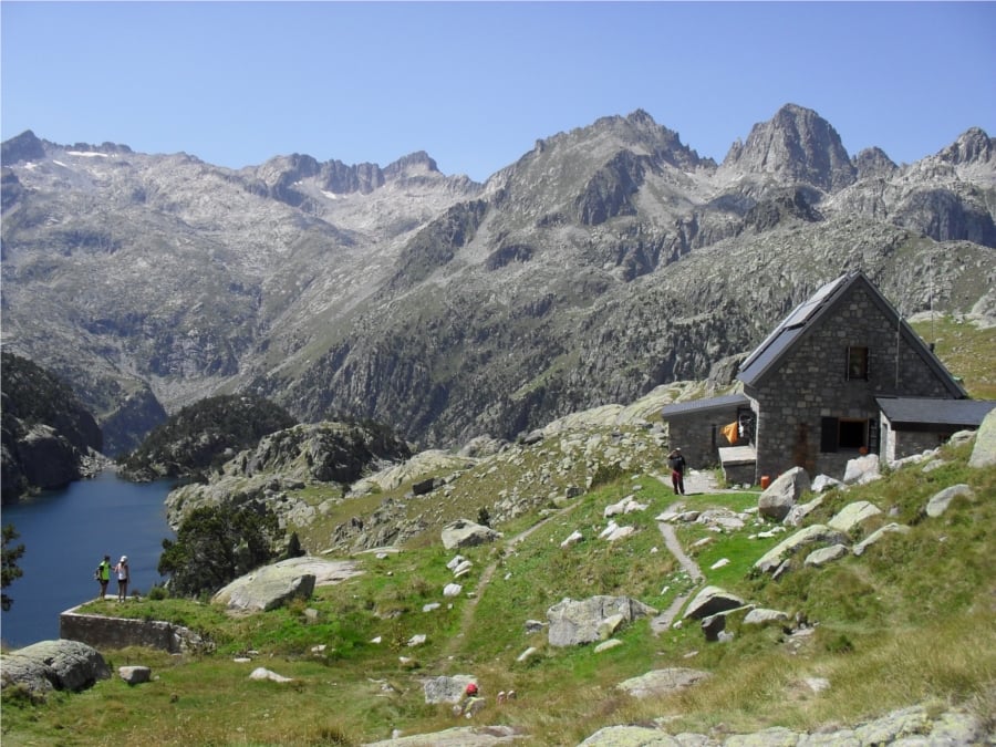 Stone mountain refuge near alpine lake with hikers and rugged peaks under clear blue sky.