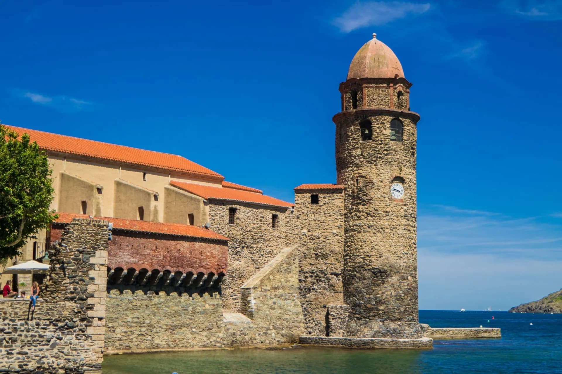 Stone church tower with red roof next to the sea under a bright blue sky in Collioure