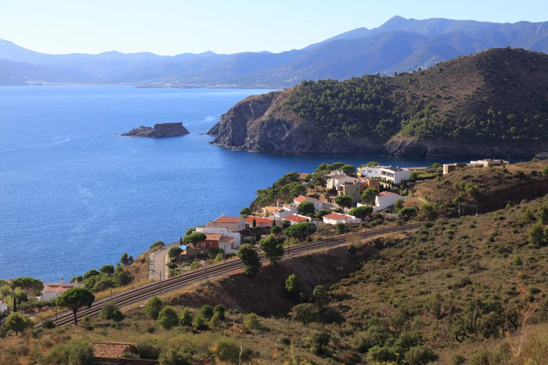 Coastal town on hillside above blue sea with mountains and railway line, Colera.