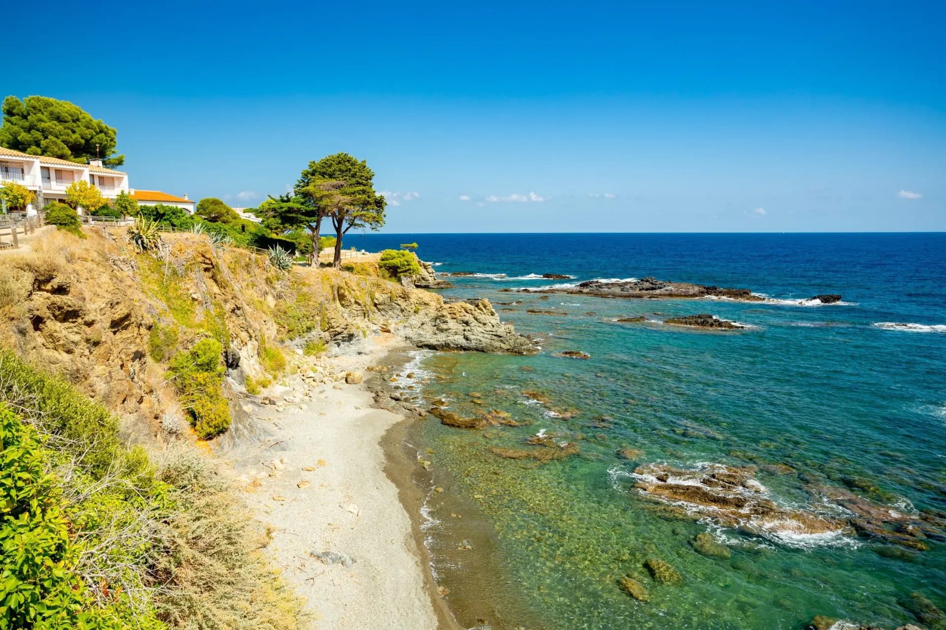 Rocky cove with clear turquoise water, dry cliffs, and white buildings near Llançà and Port de la Selva.