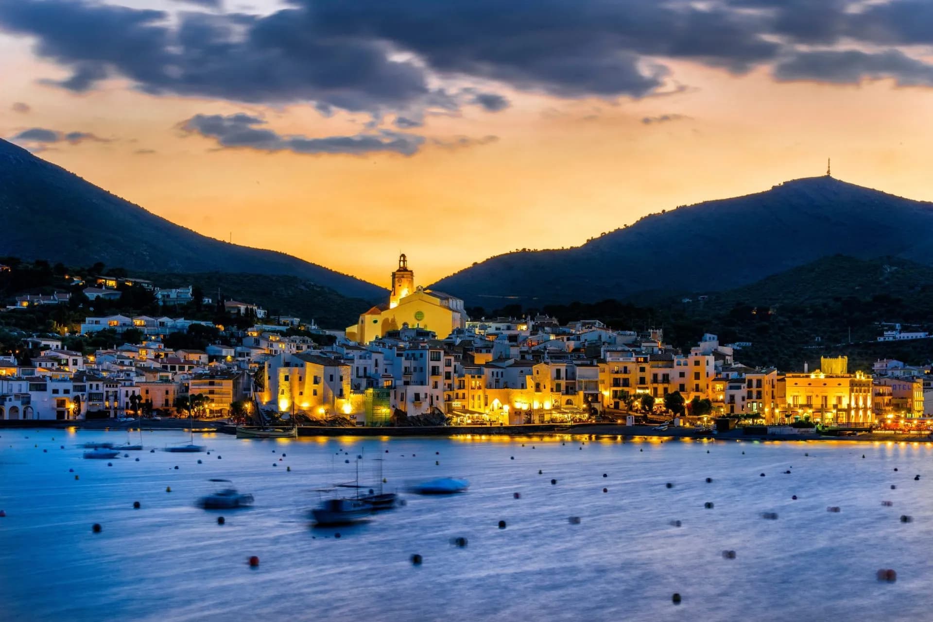 Coastal town of Cadaques illuminated at sunset with boats on the water and mountains.
