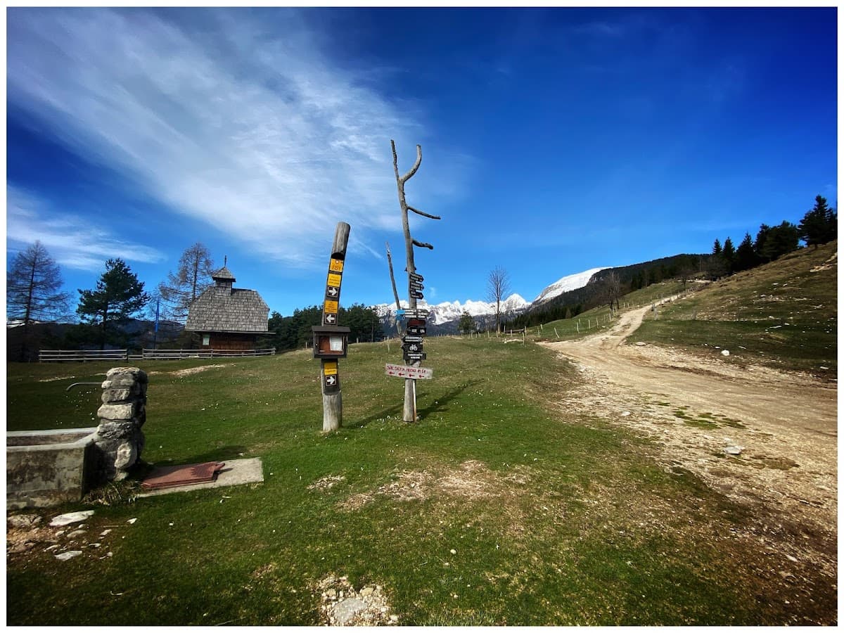 Wooden signpost on grassy mountain slope near chapel with snowy peaks under blue sky