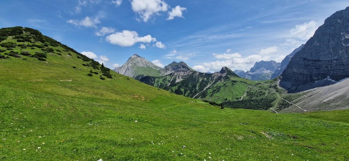 Vast green alpine meadow sloping toward rugged mountains under a blue sky with white clouds.