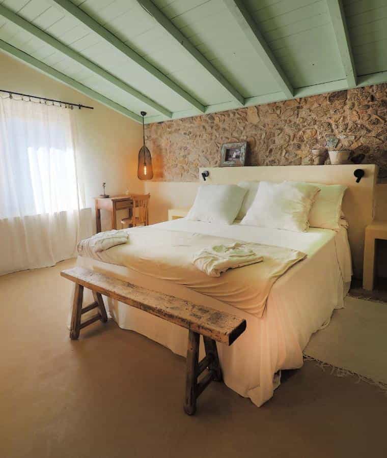 Bedroom with white linens, rustic wood bench, stone accent wall, and pale green ceiling beams.