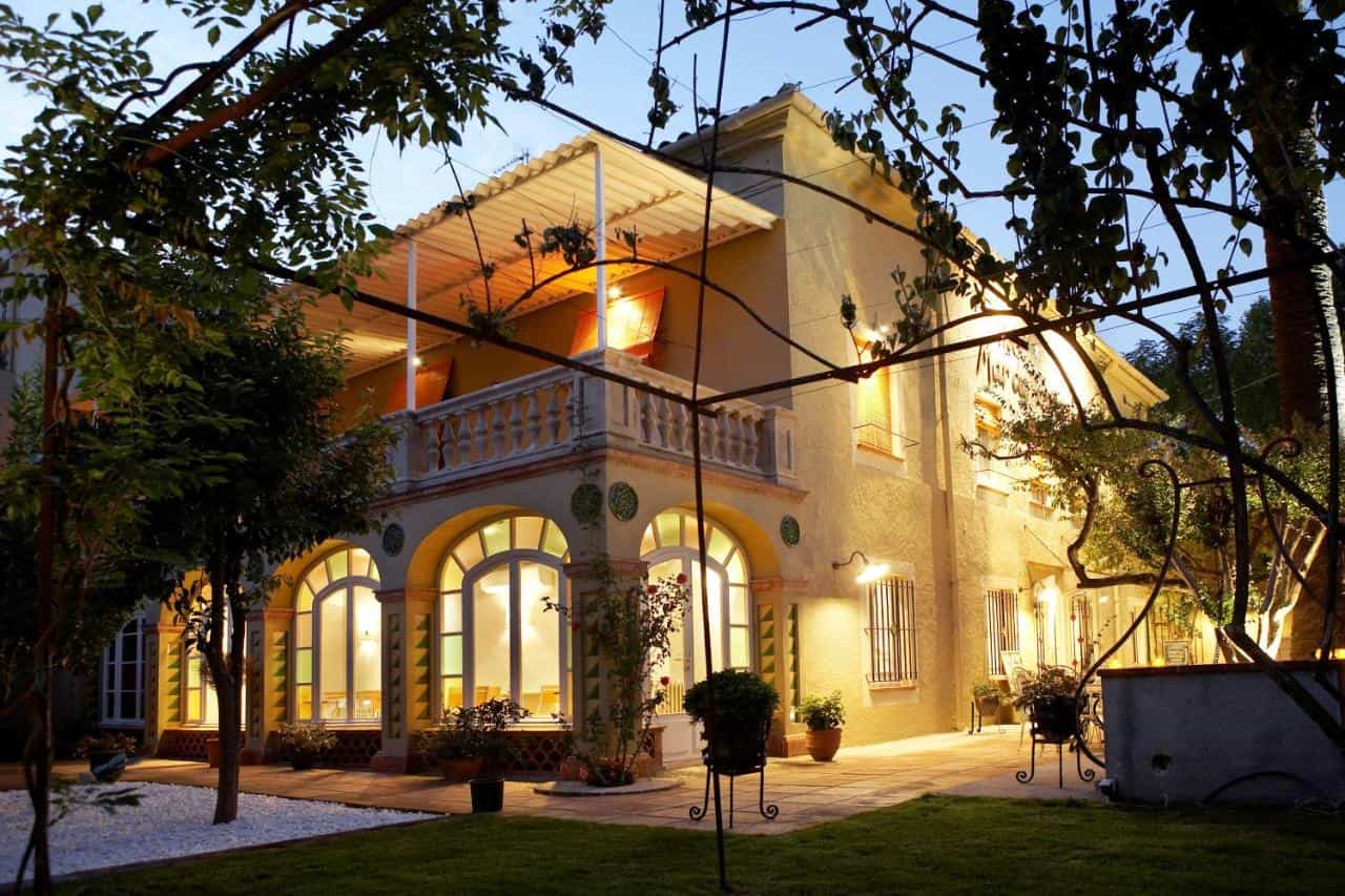 Illuminated hotel exterior with arched windows and balcony viewed through leafy garden trellis at dusk.