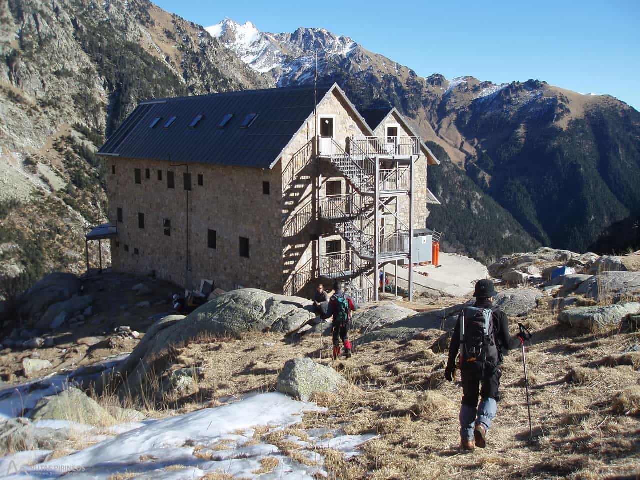 Hikers approach Refugio Ángel Orús stone mountain refuge with snowy peaks in background