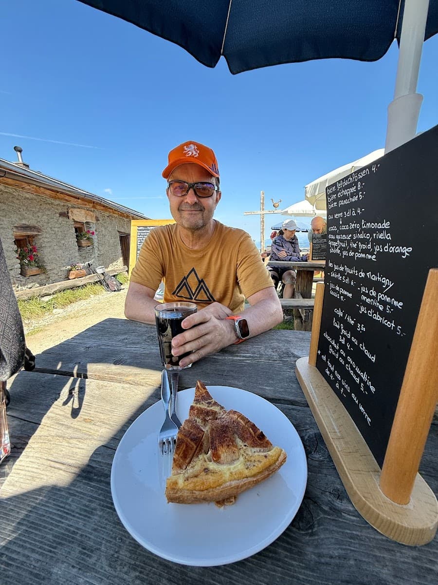 Man with orange cap dining outdoors with slice of tart and dark drink near stone building.