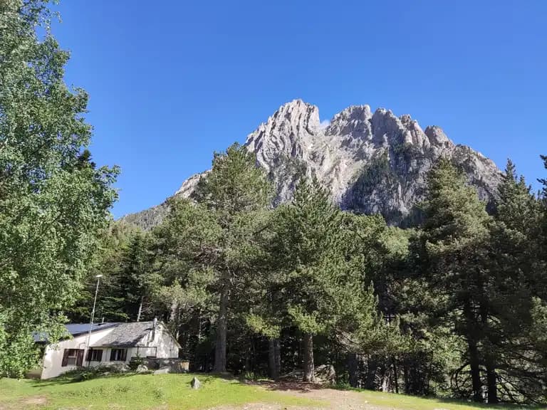 Mountain refuge hut nestled among pine trees below a rugged, rocky peak under a clear blue sky.