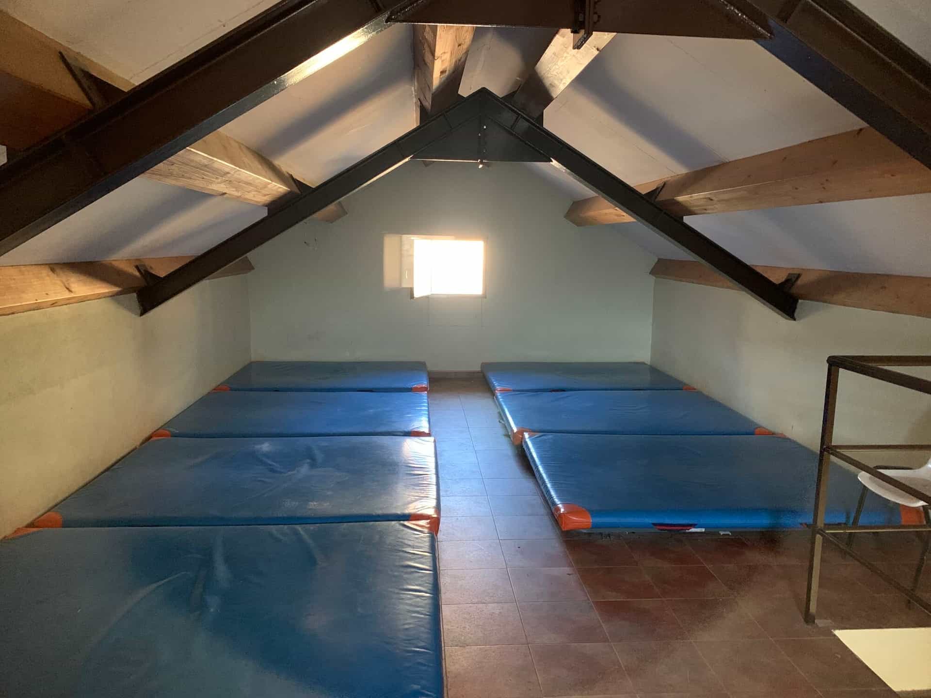 Attic dormitory with blue sleeping mats, exposed wooden beams, and a bright window at Refugio Viadós.