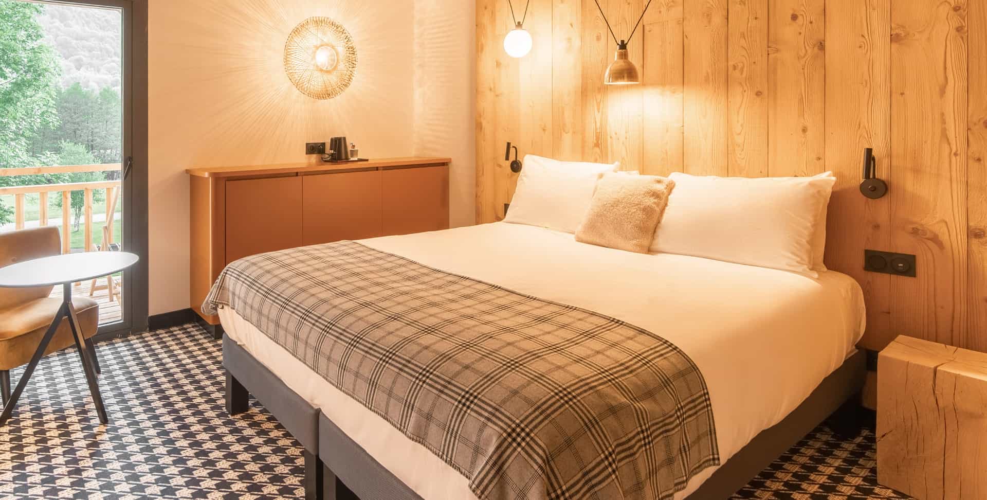 Hotel room with wood paneling, plaid throw, and balcony view of green mountains in Loudenvielle-Pyrenees.