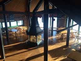 Interior of a rustic wooden lodge with a central fireplace and dining tables.