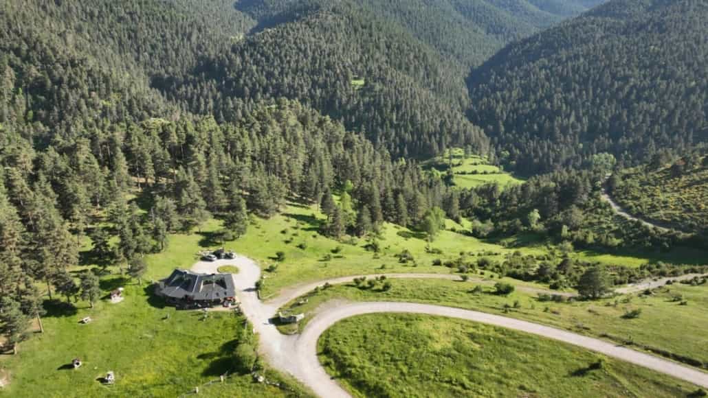 Mountain lodge nestled in green valley with winding dirt road and dense pine forests, Serrat de les Esposes