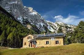 Stone mountain refuge with solar panels nestled below snow-capped peaks and pine forest.