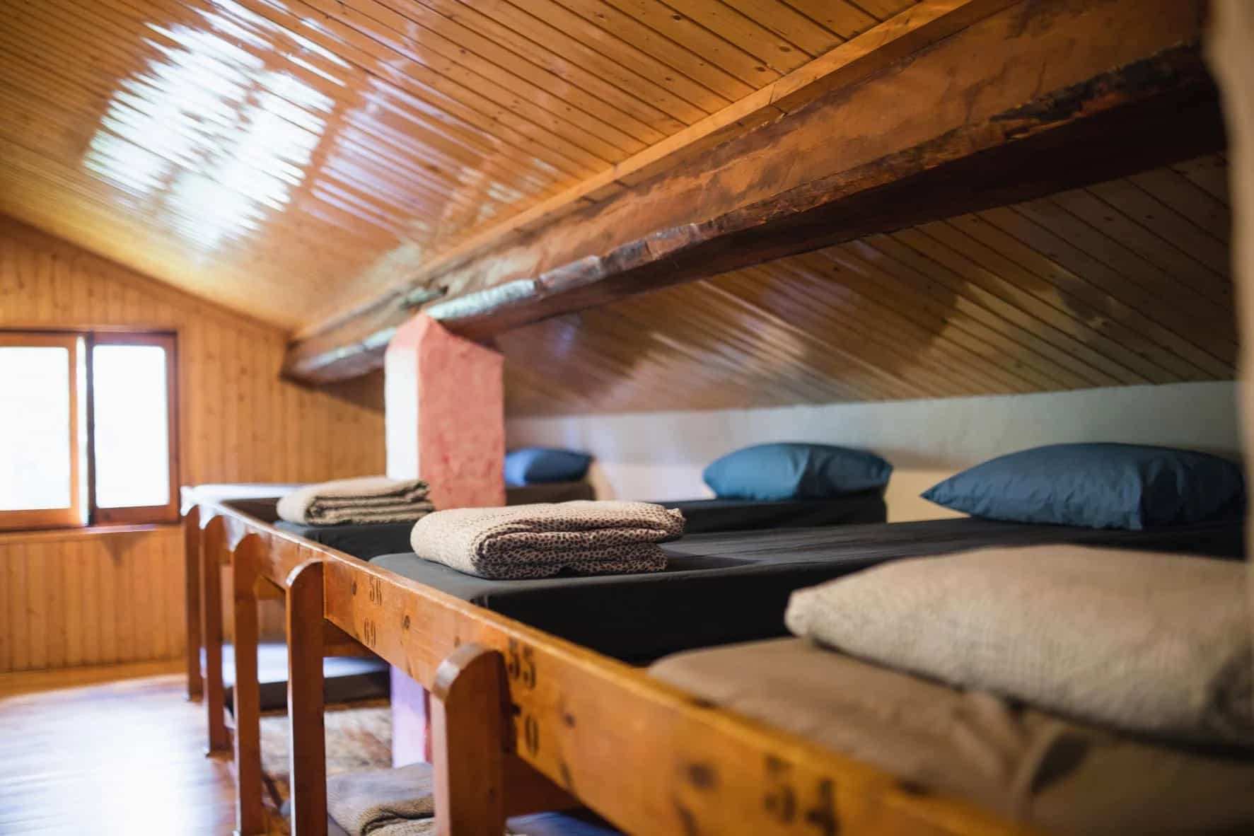 Bunk beds with folded blankets and blue pillows inside a wooden attic room with a window