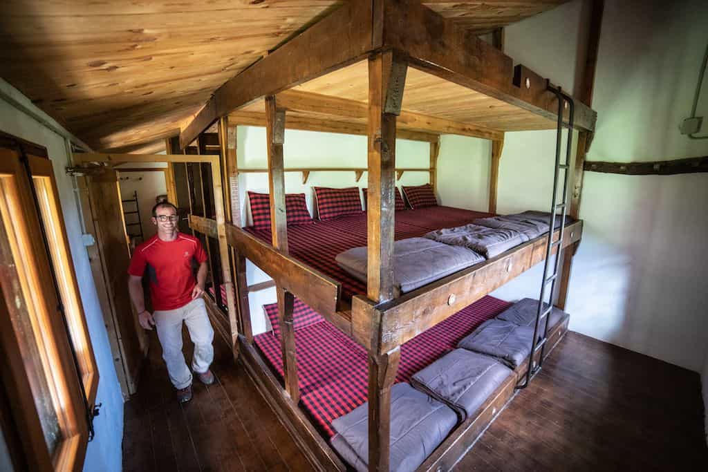 Man standing next to large wooden bunk beds with red plaid bedding inside a rustic room.