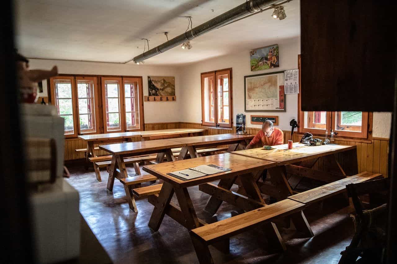 Interior of a mountain refuge dining hall with wooden tables and benches, one person seated.
