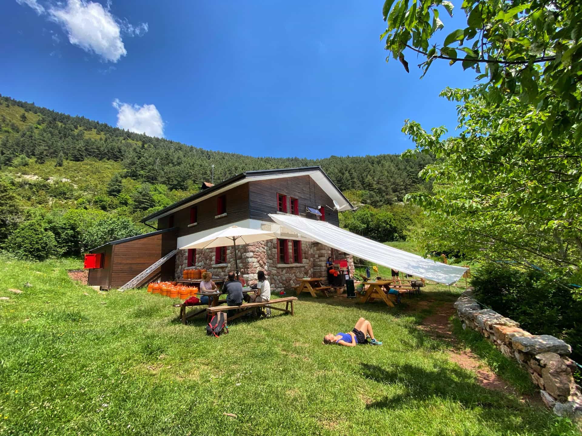 Mountain refuge Refugi Sant Jordi with people relaxing on grassy area under blue sky.