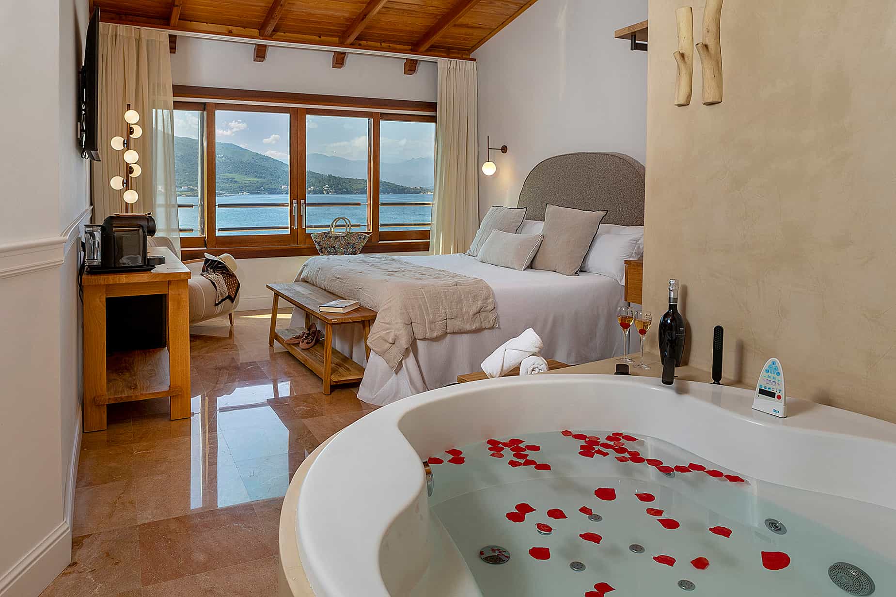 Hotel room with jacuzzi, rose petals, wine, and sea view of Porto Cristo mountains.